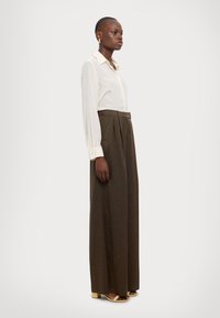 Krämsfärgad långärmad skjorta med knappar, i kombination med höga, bruna veckade byxor. Modellen har på sig sandaler med guldfärgade klackar.