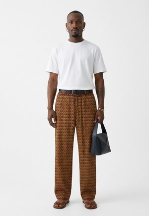 Homme portant un t-shirt blanc, un pantalon large marron à motifs, une ceinture noire, des sandales, tenant un sac à main noir géométrique sur un fond uni.