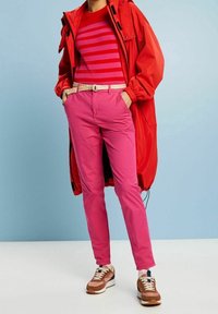 Personne portant une chemise rayée rouge et rose, une longue veste rouge vif, un pantalon rose avec ceinture tissée et des baskets marron, debout devant un fond bleu.