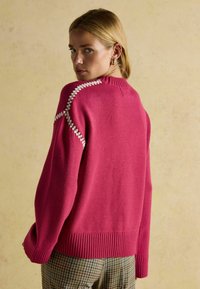 Pulover fuchsie cu tivuri cu ochiuri și cusături albe distinctive pe marginea umerilor; purtat cu pantaloni cu carouri.