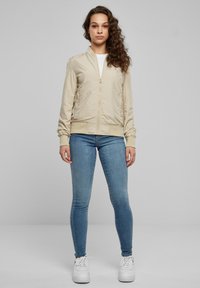 Bézs bomber dzseki cipzáros elülső résszel, bordázott szegélyekkel az ujjakon és az alján, világoskék szűk farmerral és fehér tornacipővel párosítva. Simább anyag textúra.