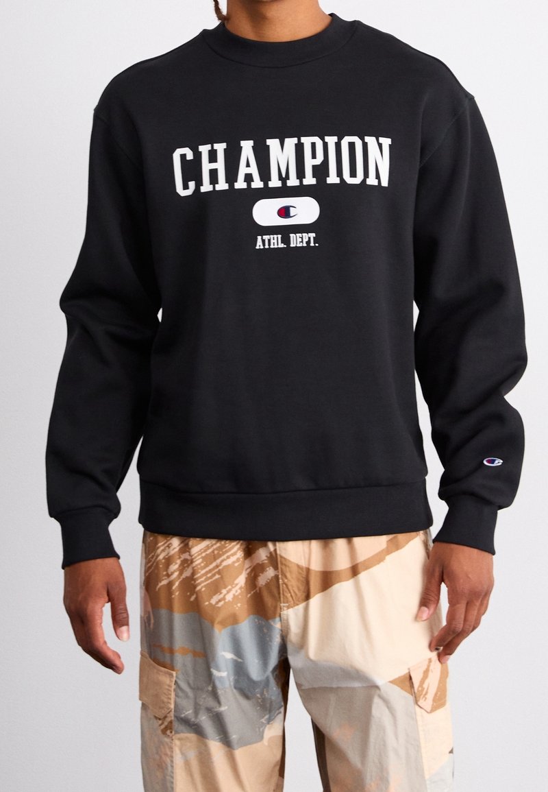 Black sweatshirt with "CHAMPION" in white text, crew neck, ribbed cuffs and hem. Worn over patterned tan and gray cargo pants.