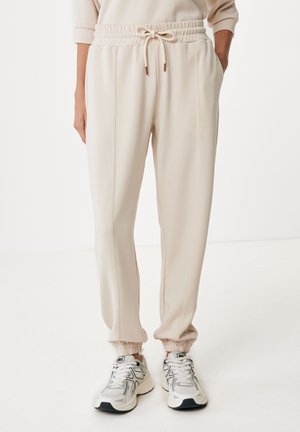 Personne portant un jogger couleur crème avec cordon de serrage, poignets élastiques et chaussures de course blanches et grises sur fond blanc uni.
