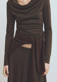 Top lung cu maneci lungi de culoare marron, cu decolteu drapat și textură plisată; detaliu legat la talie; asortat cu o fustă marron.