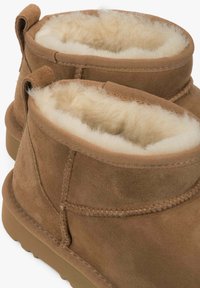 Brązowe zamszowe botki za kostkę z grubą kremową wełnianą podszewką i pętlami do ściągania z tyłu, pokazane z bliska.