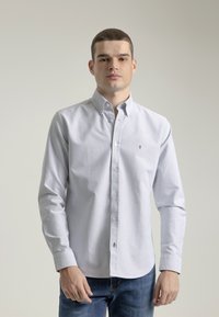 Camisa de botón en blanco con rayas verticales azules, que cuenta con un collar, mangas largas y un pequeño logo en el lado izquierdo del pecho. Tela de mezcla de algodón.