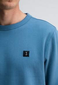 Lichtblauwe sweatshirt met een ronde halslijn en geribbelde manchetten. Heeft een klein zwart vierkant label met een witte 'J' op de linkerborst.
