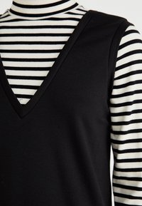 Haut noir sans manches avec un col en V superposé sur un col roulé rayé blanc et noir. Les tissus semblent doux et ajustés, créant un effet superposé.