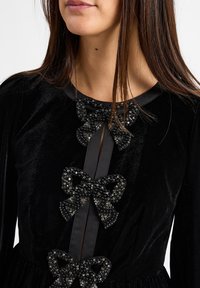 Schwarzes Samtkleid mit rundem Ausschnitt, verziert mit drei großen, gefädelten Schleifen in der Mitte und einem Detail aus schwarzem Satinband.