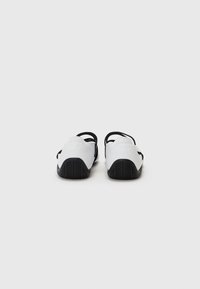 Baskets blanches sportives avec des sangles noires et des semelles en caoutchouc. Elles présentent un motif de semelle texturé et un design épuré. Style minimaliste, sans marque visible.