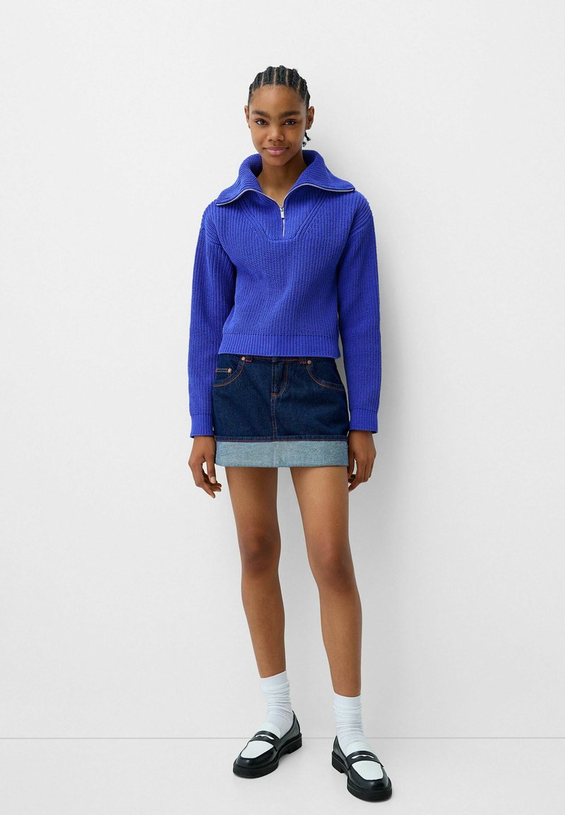 Maglione lavorato a maglia viola con colletto a zip, abbinato a una mini gonna in denim con orlo azzurro. La modella indossa calzini bianchi e scarpe nere.