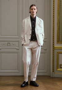 Ljusbeige kostym med en skräddarsydd kavaj, kombinerad med en svart skjorta med prickar och svarta skor. Mjuk textur, modern passform och minimalistisk design.