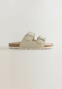 Beige Sandale mit zwei Riemen, metallischen Schnallen und dekorativen Nietenakzenten. Verfügt über ein Korkfußbett und eine strukturierte Sohle für besseren Halt.