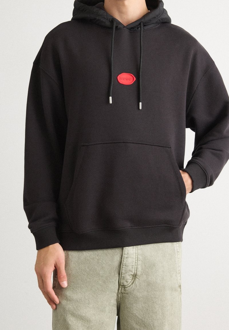 Personne portant un sweat à capuche noir avec un patch logo rouge « HUGO » sur la poitrine, pantalon beige clair, une main dans la poche, devant un mur uni.