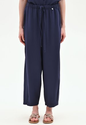 Pantaloni larghi blu navy con vita regolabile con cordoncino e tasche laterali, indossati con sandali bassi intrecciati beige su una persona su sfondo bianco.