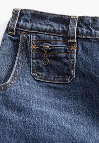 Denim jeans featuring a dark blue shade, with contrasting orange stitching. Notable details include a small zippered pocket and sturdy waistband.