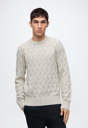 Man wearing a light beige cable-knit sweater with ribbed cuffs and hem, standing against a plain white background.