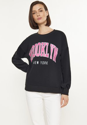 Fekete kapucnis pulóver, oversize kialakítással, rózsaszín "BROOKLYN" felirattal, bordázott mandzsettákkal és aljjal. Sima textúra, fehér nadrággal viselve.