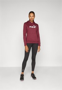 Maroon dukserica s kapuljačom i prednjim džepom, s bijelim logotipom, u kombinaciji s crnim tajicama sa bočnim detaljima i crnim sportskim cipelama.