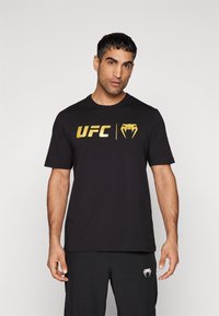 Sort T-shirt lavet af bomuld med et stort guld UFC-logo og en slange grafik. Rund hals og korte ærmer, med en glat tekstur.