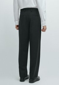 Schwarze Hose mit geradem Bein, glatter Textur, Gürtelschlaufen und zwei Gesäßtaschen. Kombiniert mit einem weißen Langarmhemd.