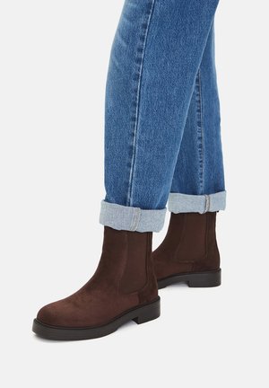 Braune Wildleder-Stiefeletten mit elastischen Seitenteilen und einer strukturierten Gummisohle, kombiniert mit hochgekrempelten blauen Jeans.