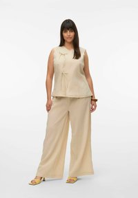 Beige, mouwloos topje met strikaccenten, gecombineerd met losse, wijde broek. Textiel met een natuurlijke afwerking. Gele sandalen maken de look compleet.