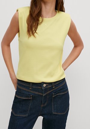 Gelbes, geripptes, ärmelloses Oberteil mit Rundhalsausschnitt, kombiniert mit dunkelblauen Jeans aus Denim, die über Fronttaschen und kontrastierende Nähte verfügen.