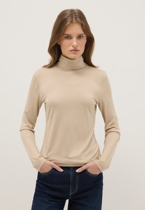 Joven con el pelo largo y castaño, vestida con un jersey de cuello alto beige y vaqueros azul oscuro, de pie con las manos en los bolsillos sobre un fondo liso.