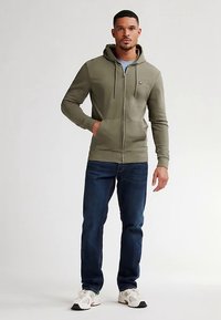 Sudadera verde con cremallera y bolsillos tipo canguro, combinada con vaqueros azules y zapatillas blancas. Textura suave, ajuste regular y capucha con cordón ajustable.