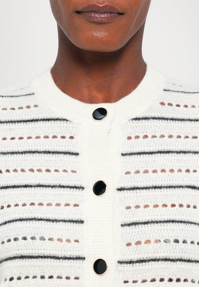Witte cardigan met zwarte horizontale strepen, met een ronde halslijn en vier grote zwarte knopen. Textured breimateriaal met uitsnijdingen.