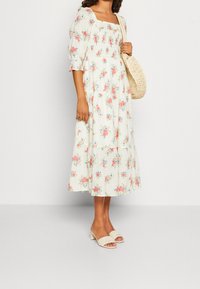 Vestido midi floral en crema con patrones en rosa y azul, escote cuadrado, mangas abullonadas y falda con volantes. Llevado con sandalias de tacón bloque en beige.
