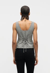 Άτομο με πλεγμένα μαλλιά φοράει αμάνικο τοπ σε ασπρόμαυρο καρό μοτίβο με κορδόνια στο πίσω μέρος, συνδυασμένο με μαύρο παντελόνι, με την πλάτη στραμμένη προς τα πίσω.