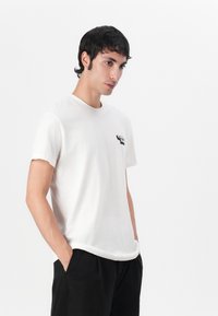 White cotton t-shirt with short sleeves, round neckline, and a small black graphic design on the left chest. Smooth texture.