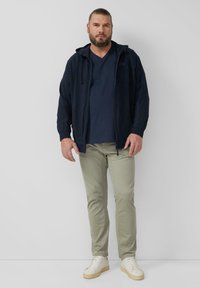 Marineblaue Kapuzenjacke mit Reißverschluss und Kordeln über einem marineblauen V-Ausschnitt-Shirt, kombiniert mit hellgrünen Hosen und cremefarbenen Sneakern mit beigefarbenen Sohlen.
