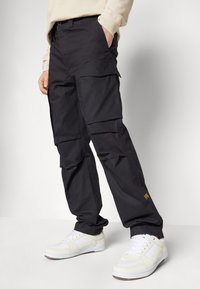 Schwarze Cargo-Hosen mit mehreren Taschen, lockerer Passform und gewebtem Material, kombiniert mit weißen Sneakern. Minimal sichtbares Branding.