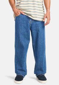 Weite blaue Jeans aus Denim mit einer leicht verblassten Textur, ausgestattet mit einem klassischen Fünf-Taschen-Design. Kombiniert mit dunklen Schuhen.