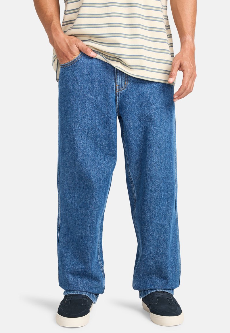 Weite blaue Jeans aus Denim mit einer leicht verblassten Textur, ausgestattet mit einem klassischen Fünf-Taschen-Design. Kombiniert mit dunklen Schuhen.