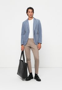 Man wearing blue blazer, white shirt, beige pants, black shoes, holding a large black tote bag against a plain white background.