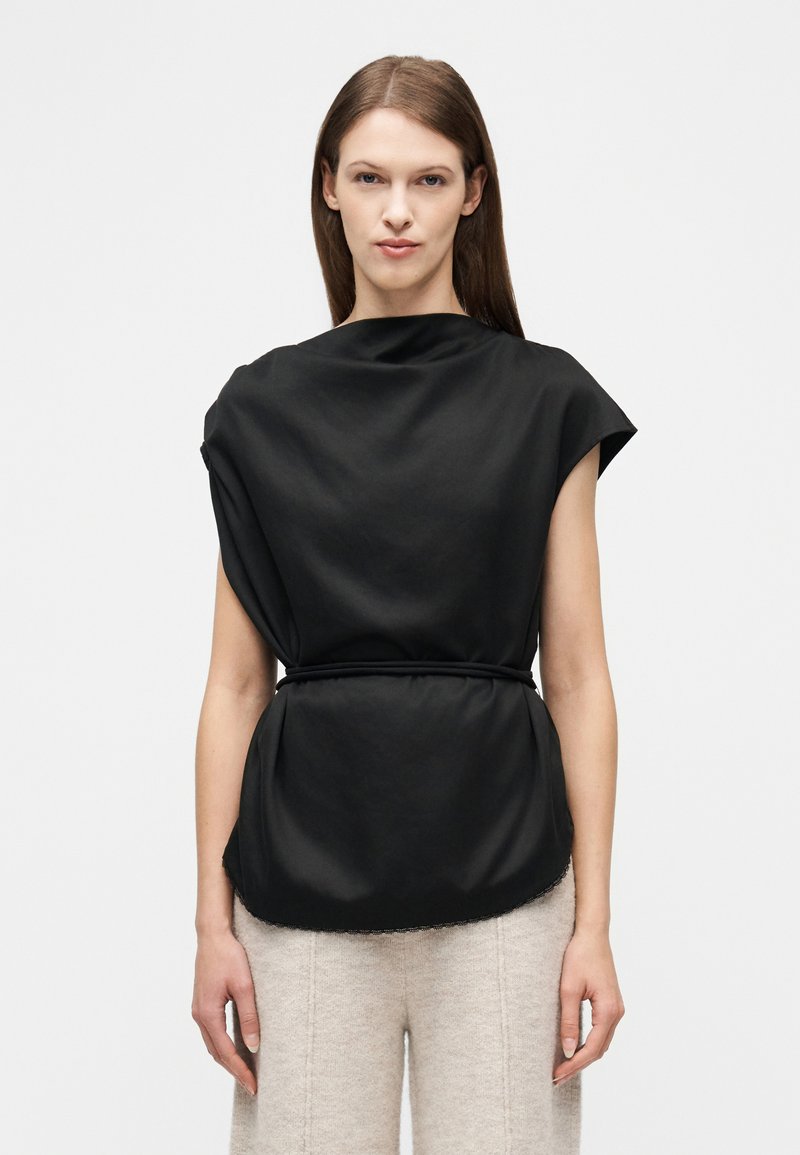 Γυναίκα με μακριά καστανά μαλλιά φοράει μαύρο αμάνικο τοπ με ζώνη και μπεζ υφασμάτινο παντελόνι, στέκεται μπροστά σε απλό φωτεινό φόντο.