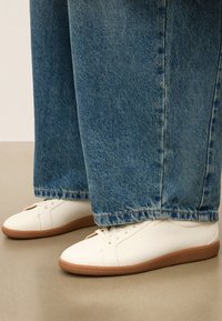 Witte lage sneakers met gumzolen, gedragen met brede blauwe jeans op een beige vloer.