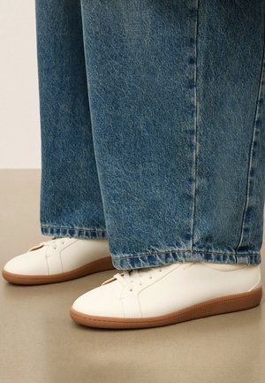 Witte lage sneakers met gumzolen, gedragen met brede blauwe jeans op een beige vloer.