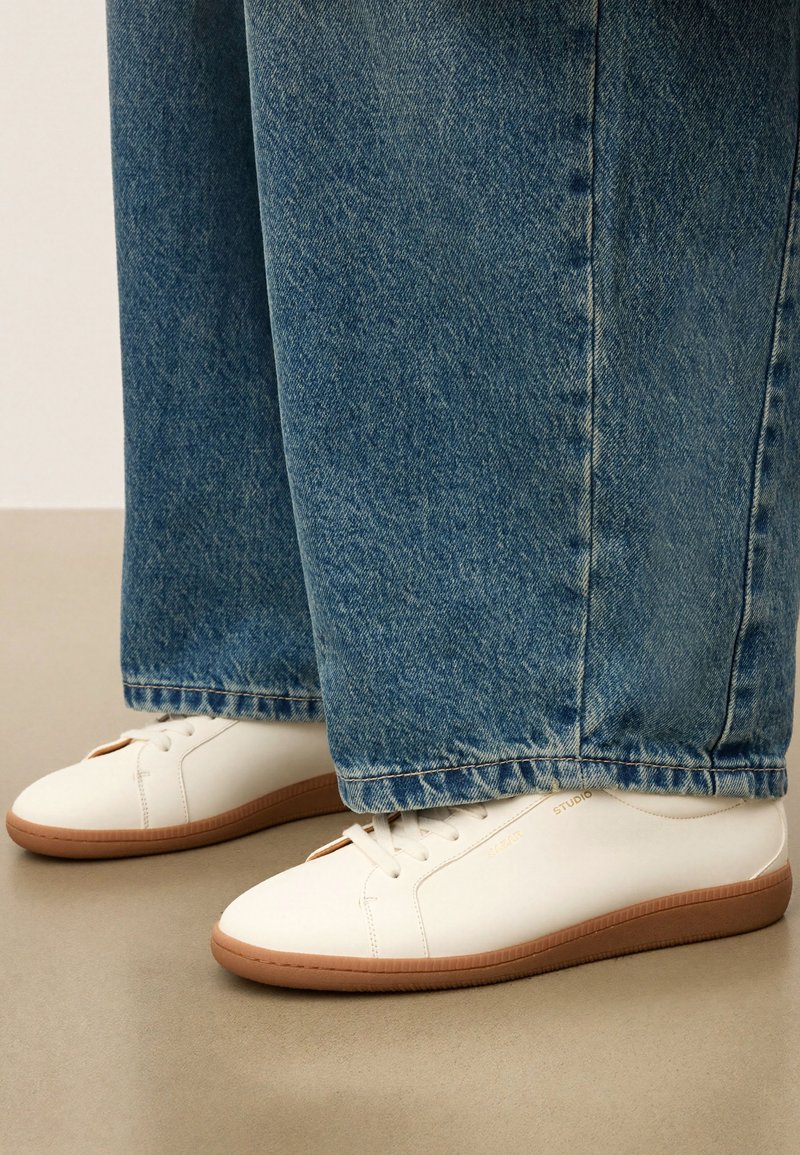 Witte lage sneakers met gumzolen, gedragen met brede blauwe jeans op een beige vloer.