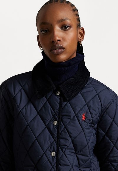 Die marineblaue gesteppte Jacke verfügt über einen Cordkragen, Metalldruckknöpfe und ein rotes gesticktes Logo auf der Brust. Glatte Textur.