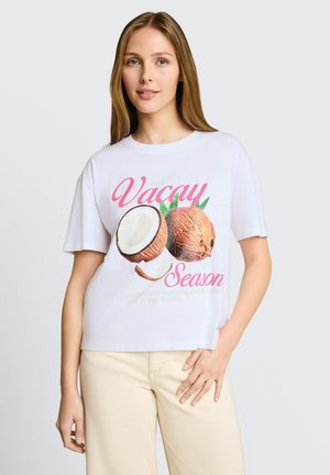 Junge Frau mit langen braunen Haaren, die ein weißes T-Shirt mit dem Text „Vacay Season“ und einem Kokosnuss-Design trägt, kombiniert mit hellbeigen Hosen.