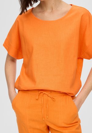 Frau trägt ein locker sitzendes, leuchtend orangefarbenes Kurzarmoberteil und passende Hosen mit Kordelzug, Hände in den Taschen.