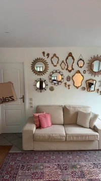Divano beige con due cuscini decorativi, circondato da vari specchi ornati con cornici dorate e argentate, su una parete bianca. Sotto, un tappeto rosa con motivi.