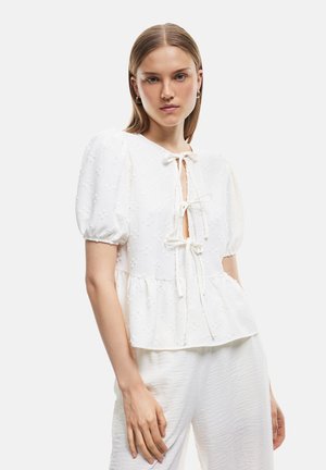 Blusa bianca a peplo con maniche a palloncino, chiusura a fiocco al collo, texture floreale e vita leggermente arricciata. Abbinata a pantaloni leggeri e testurizzati.