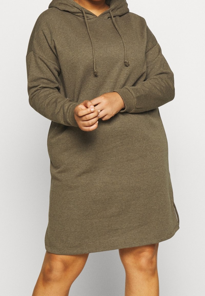 Personne portant une robe sweat à capuche vert olive à manches longues avec des cordons, debout les mains jointes et les jambes visibles sous l'ourlet de la robe.