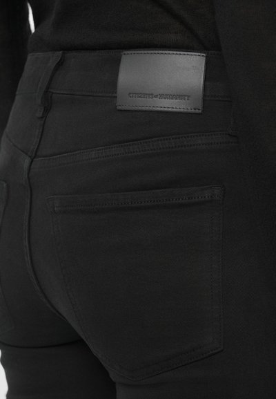 Μαύρο τζιν από τζιν ύφασμα με δερμάτινο λογότυπο στη ζώνη, με κοντινή εικόνα στις πίσω τσέπες και αντιθετικές ραφές.